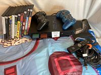 Photo showing two PlayStation 2 consoles with two controllers (black and blue translucent), a remote control, and the 10 games lined up vertically along with one dance pad visible.