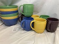 Full view of 6 stacked ceramic bowls in green, yellow, and blue, and 7 ceramic mugs with handles in green, yellow, blue, and maroon colors