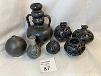 Seven black pottery vessels shown together, highlighting variety of shapes and sizes including a tall double-handled vessel and smaller spherical vessels with cut-out details.