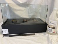 Front view of the ethanol tabletop fireplace showing three flame burners with black base and glass enclosure, a bottle of ethanol fuel placed beside it, ruler for scale.