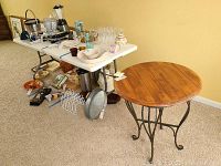 White folding table with items on top and some scuff marks on legs, with round wood and metal occasional table to the side