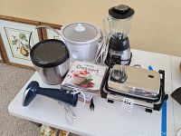 Photo showing all items in the lot including blender, crock pot, panini press, ice bucket, steamer, and cookbooks on white table.