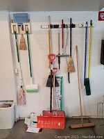 Wide shot showing 9 items including plastic snow shovels, mops with green and pink heads, several brooms hung on wall in garage