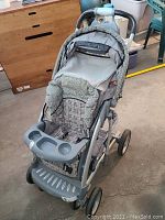Front and side view showing the fabric seat with plaid insert, child's gray plastic tray with compartments, padded handlebars, and canopy of the stroller.