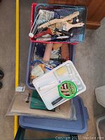 View of stacked plastic paint trays in red, blue, and white colors with painting tools inside and various rolls of tape on top.