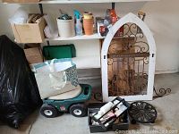Wide view of lot showing decorative door, plastic gardening pots, garden wagon, cardboard boxes, first aid kit storage boxes, and shelves with garden supplies.