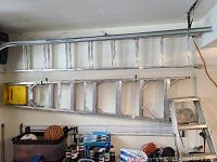 Three aluminum ladders: one extension ladder and two step ladders arranged along a wall in a garage.