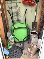 Wide view of green wheelbarrow, tomato fertilizer, metal umbrella stand base, and some garden supplies stored in shed