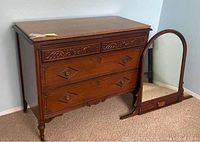 Antique 3 drawer wooden chest with granite top next to a wooden framed standing mirror with rounded top.