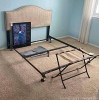 Full view of queen bed frame with beige upholstered headboard, metal bed frame, adjacent luggage rack, framed print leaning against the frame, and under-bed storage bags on carpeted floor in bedroom.