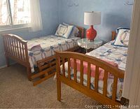 Photo showing two separate twin wooden beds with patchwork quilts and pillows, rails leaning on the floor between beds, lamp and table present.