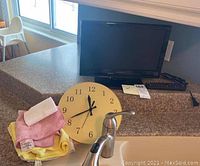 16 inch Dynex flat screen TV placed on a kitchen counter with remote control beside it, a yellow and pink kitchen towel set in front, and a round kitchen clock behind the towels.