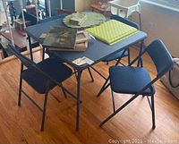 Blue folding card table with 4 matching blue padded folding chairs. Table is square with metal legs and padded top. Chairs have metal frames and padded seats and backs with quilted texture upholstery.