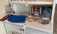 Photo of entire group showing various silver-plated serving pieces and flatware arranged on white cabinet.
