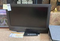 Full frontal view of the 27-inch Olevia flat-screen LCD TV standing on carpet, with the remote and instruction sheet placed in front.