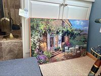 Large canvas wall art with outdoor cottage scene featuring a table with two chairs and flowering vines above entrance.