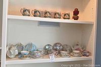 Shelves containing 12 delicate teacups and saucers with mixed floral and gold trim designs from various countries, some on metal display stands.
