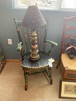Full view of hand painted wooden arm chair with table lamp placed on the seat. Chair features black base color with gold dots and floral detail, green armrests, and decorative turned legs.