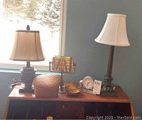 Photo of all items showing three lamps, brass bowl, woven basket with lid, and quartz clock on a wooden cabinet.