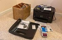 Wide shot of Epson WF-2860 printer with paper cutter, label maker, manuals and bag of file folders on carpeted floor.