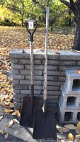 Photo showing two metal shovels placed side by side against a brick wall outdoors. One shovel has a D-shaped grip wood handle, the other has a straight wood handle.