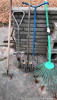 Photo showing five garden tools including a green plastic leaf rake, a garden hoe, a hand rake, a hand cultivator, and a stainless steel bulb planter.