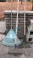 Three garden rakes standing upright against a stone wall; includes one plastic green rake, one metal rake, and one smaller metal rake.