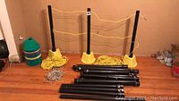 Overview photo showing 24 yellow plastic bases, 19 black plastic conduit poles, and coiled yellow nylon rope on hardwood floor against a wall.