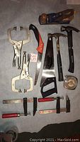 Top view of miscellaneous hand tools arranged on floor including two hammers, two saws, four clamps, chalk line tool, wood hand lathe tool, and measuring tape.