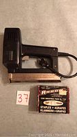Black electric staple gun with trigger handle and attached power cord paired with a box of Arrow industrial staples. Vendor tag numbered '37' visible next to items.
