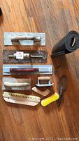 Overview photo of the assorted cement finishing tools arranged on wooden floor including floats, brushes, trowels, and a rolled mat.