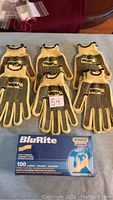 Six pairs of yellow and black cotton gloves with rubber grip dots on the palms displayed flat; box of blue nitrile gloves in front.