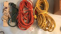 Overview of the lot showing two coiled long extension cords (orange and yellow) and two power bars (beige and green) on a light surface with a lot number card '69'.