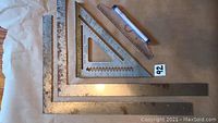 Assorted metal squares and angles including large carpenter's square, metal triangular square, and a small clear plastic protractor ruler with bubble level.