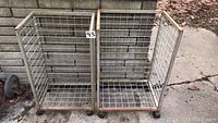 Two rectangular metal wire mesh carts side by side on a concrete surface, showing open tops and grid pattern. Both have rust spots and caster wheels visible.