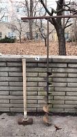 Full view of hand auger and sledgehammer leaning against a brick wall outdoors with leaf litter on the ground.