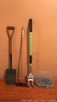 Photo showing shovel, long-handled magnet sweeper, metal roofing shingle remover, and plastic container of nails on wooden floor against a plain background.