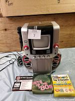 Front view of the karaoke machine with built-in CRT screen, red speakers, microphone, control knobs, and karaoke CD sets in front.