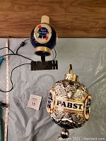 Two Pabst Blue Ribbon-themed light fixtures displayed on a table, showing the table lamp with blue logo globe and the wall-mount lamp with etched glass globe.