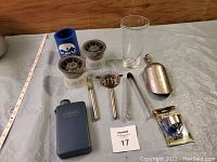 Photo showing two round ice ball molds, cocktail strainer, tongs, jigger, blue rubber bottle opener, metal flask, and a blue cylindrical flask.