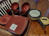 Photo showing 4 large square plates, 3 small plates, 3 cups, 4 bowls, and a saucepan with lid on wooden table