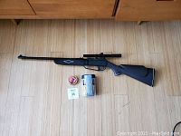 Photo showing the Daisy pellet rifle with mounted scope, round container of pellets, and target pack on wooden floor.