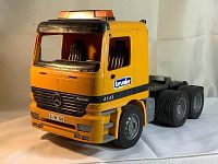 Front view showing yellow truck cab with Bruder branding and Mercedes emblem, black grille, orange light bar on rooftop, and missing windshield.