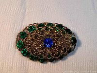 Front view of the brooch pin showing sapphire blue center stone and surrounding emerald green rhinestones with intricate copper filigree design.