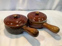 Two brown pottery French onion soup crocks with lids and handles, side-by-side on white surface