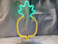 LED pineapple light sign turned on, showing yellow light on the pineapple body and green light on the leaves, on a white round base.