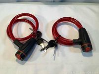 Two new red bike lock cables with attached keys laid side by side on a white surface, showing overall condition and length.