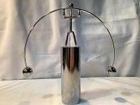 Front view of metallic kinetic weightlifter desk toy showing a weightlifter figure balanced on a curved bar with spherical weights at the ends, mounted on a cylindrical base.