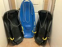 Three plastic sleds arranged against a wall showing two black Torpedo sleds with yellow brake handles on sides and a blue Pelican sled in the center with attached ropes.