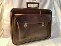 Front angled view of the soft brown leather briefcase showing the flap pocket with two metal push-button clasps and the main handle on top. A key is attached to the lock mechanism.